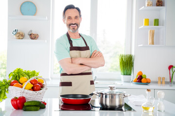 Photo of charming confident mature guy wear blue t-shirt apron cooking arms crossed smiling indoors house room
