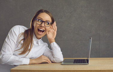 Young woman sitting near a laptop trying to eavesdrop on something by putting her hand to her ear....