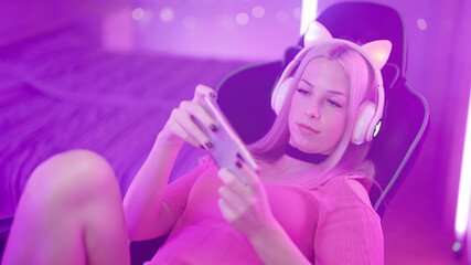 Fototapeta na wymiar Cute Blonde Gamer Girl on Gaming Chair playing Games on Smartphone