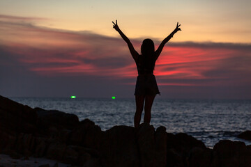 Fototapeta premium Woman standing waving on the rocks by the sea in the red sky at twilight at Patong Beach, Phuket, Thailand..