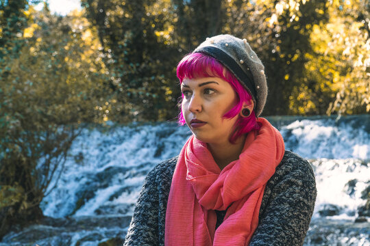 Mujer con gorro gris y bufanda rosa delante de una peque&ntilde;a cascada, mujer de pelo rosa mirando hacia la izquierda , mujer seria pensando junto al r&iacute;o , mujer con ojos claros y labios rojos melanc&oacute;lica