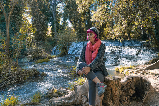 Mujer con bufanda rosa sentada en un tronco de madera delante de la cascada, mujer pensativa con las piernas cruzadas junto al r&iacute;o, chica con gorro y jersey gris sentada junto al r&iacute;o  