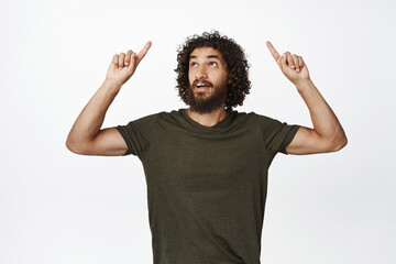 Image of young curly guy pointing fingers up, looking amazed by sale offer, standing in t-shirt...