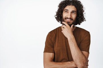 Fototapeta na wymiar Portrait of smiling curly man thinking, imaging smth, touching his beard with thoughtful face expression, choosing, standing over white background