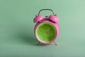 Tasty matcha tea in a pink retro alarm clock on green background. Waking up with alarm and green...