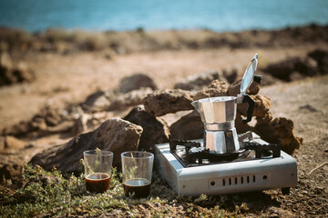 Outdoor coffee preparation with the moka pot on the camping stove on the beach with the ocean in the background, brewing espresso, glasses