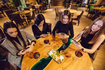 A group of women friends are sitting at a table in a restaurant and using smartphones.