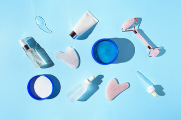Cosmetic facial kit for home skin care and spa. Collagen eye patches, face roller and gua sha massagers, bottles of essential oils and serum on blue pastel sunlit background. 