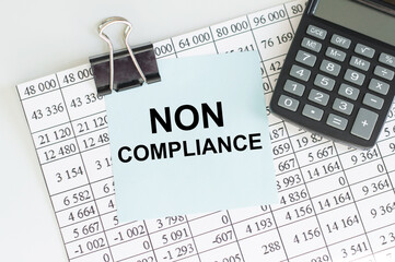 text NON COMPLIANCE in the blue short note paper on the background of a sheet with numbers next to a pen and a calculator on the table