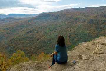 Rear view of a beautiful woman, adventurer or tourist, resting on the edge of a cliff overlooking the autumn mountains.