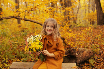 a beautiful blonde girl in a mustard coat is sitting on a bench in the autumn forest with a bouquet of flowers