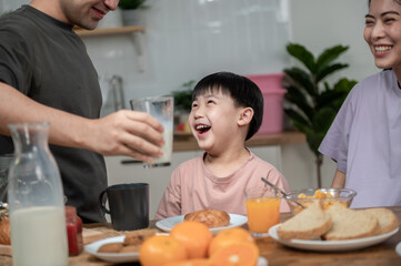 Obraz na płótnie Canvas Happy Asian family enjoying breakfast together on dinning table. Young Asian father taking a glass of milk to his son while having breakfast in the kitchen. Happy Asian family lifestyle concept.
