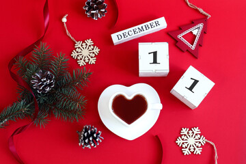 Calendar for December 11: the name of the month in English, cubes with the number 11, a cup of tea in the shape of a heart, a fir branch, cones, Christmas decor on a red background, top view