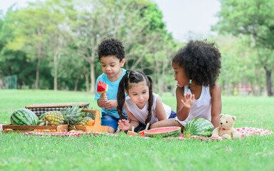 Three mixed race caucasian little cute children kids sitting, playing in outdoor green park for picnic, eating fruit, watermelon and pineapple with freshness. Education, Friendship, Diversity Concept.