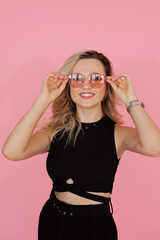 Portrait of charming blonde in sunglasses on pink background in studio. young woman smiles, looks happily into frame. dressed in a black dress with ribbons crossed on her stomach.
