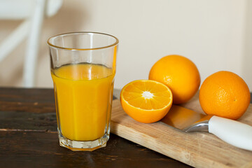 A glass of orange juice and fresh oranges and cutting board with a knife are on a wooden table in the kitchen.