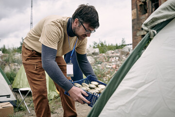 Male volunteer with box of canned food bending by tent where migrants live