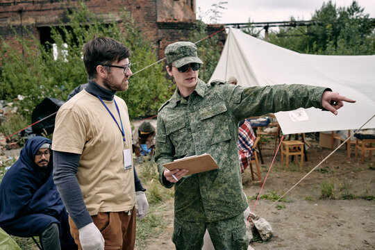 Young Volunteers Discussing Improvement Measures In Refugee Camp Against Tent And Group Of Migrants