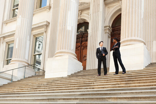 A well dressed man and woman converse on the steps of a legal or municipal building. Could be business or legal professionals or lawyer and client.