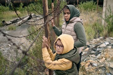Two little girls in hoodies and jackets standing by net dividing zone of refugee camp from other...
