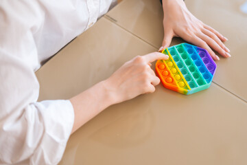 Close-up hands of unrecognizable young woman playing with rainbow pop-it fidget toy sitting at...