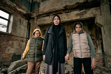 Fototapeta na wymiar Young Muslim woman holding her two daughters by hands while standing in half ruined house