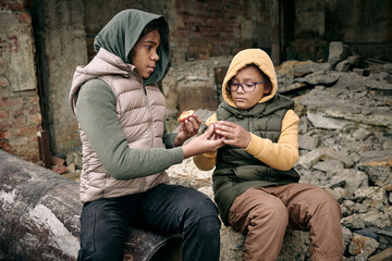 Two hungry girls from refugee camp sharing peach while sitting on ruins of building