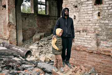 Young man and little girl standing by abandoned ruined building against brick wall