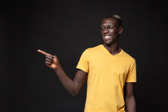 Smiling young african american man guy in yellow t-shirt posing isolated on black wall background studio portrait. People emotions lifestyle concept. Mock up copy space. Pointing index finger aside.