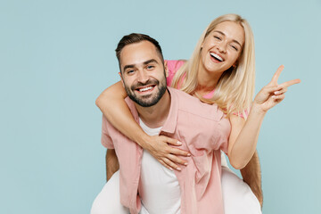 Young smiling fun couple two friends family man woman in casual clothes giving piggyback ride to joyful, sit on back show v-sign gesture together isolated on pastel plain light blue background studio.