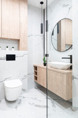 Modern bathroom interior with marble tiles, wooden furniture, glass, round mirror and stylish ceramic washbasin.  Luxury bathroom with sink and toilet. Vertical.