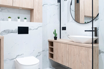 Contemporary bathroom with glass, wooden furniture, ceramic sink and marble tiles on the wall. Interior of modern bathroom with toilet and round mirror.