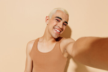 Close up young smiling happy blond latin gay man with make up in beige tank shirt doing selfie shot...