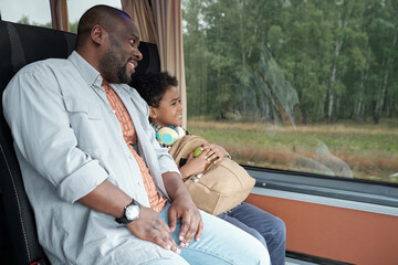 Young father and cute son with backpack looking through bus window while riding through forest