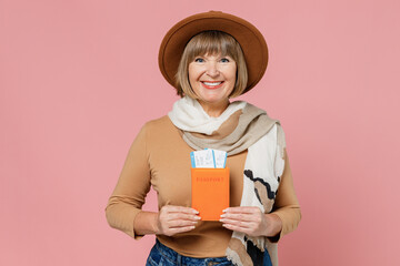 Traveler tourist happy smiling mature elderly senior lady woman 55 years old wears brown shirt hat scarf hold passport boarding tickets isolated on plain pastel light pink background studio portrait.