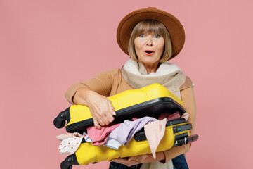 Traveler tourist surprised mature elderly senior lady woman 55 years old wears brown shirt hat scarf hold open suitcase bag with clothes isolated on plain pastel light pink background studio portrait.