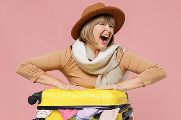 Traveler tourist mature senior woman 55 years old wears brown shirt hat scarf try to close open suitcase bag with clothes scream aside isolated on plain pastel light pink background studio portrait.