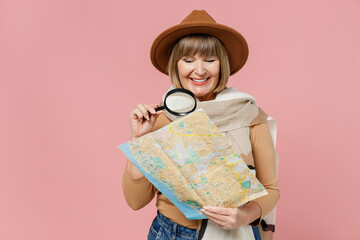 Traveler tourist excited mature elderly senior lady woman 55 years old wear brown shirt hat scarf hold examine map with magnifying glass isolated on plain pastel light pink background studio portrait