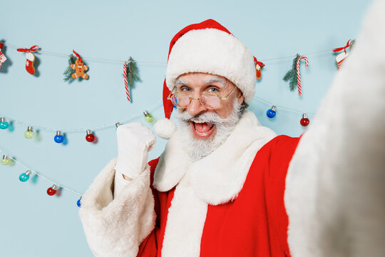 Close up old bearded Santa Claus man in Christmas hat red suit do selfie shot pov on mobile phone do winner gesture isolated on plain blue background studio Happy New Year 2022 merry ho x-mas concept.