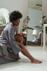 African female in grey homewear applying moisturizing cream on legs after morning shower