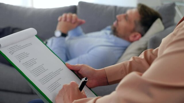 Unfocused adult man with mental health problems after covid-19 lying on couch talking to therapist. Close up woman psychologist listens patient and taking notes on clipboard at psychotherapy session