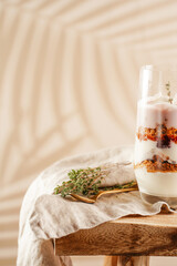 Obraz na płótnie Canvas Tall glass with a layered creamy yoghurt dessert trifle with cookies, amaretti, cherries and thyme on a linen cloth on a rustic wooden table at a tropical setting