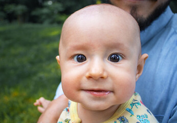 Kid looking at the camera and smiling close-up. Dad holds his baby son in his arms outdoors.
