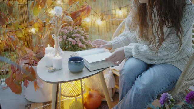 Young woman leafs through book and takes warm beverage cup sitting in armchair on balcony decorated with yellow lights and autumn attributes in evening