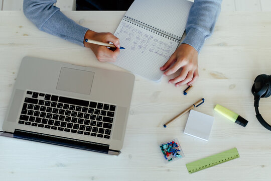 Crop student writing in notepad at desk with laptop