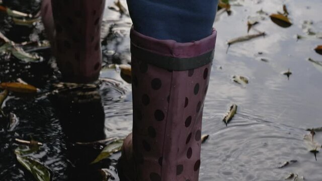 Child girl or teen in rubber boots are walking on wet asphalt covered with puddles and fallen leaves. Children and autumn. Full HD slow motion video