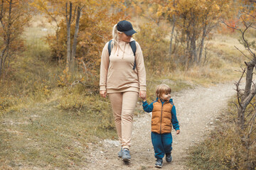 Full body portrait of caucasian happy family: mother with backpack in beige sportswear walking with her little baby boy 2-3 years old in the fall woods on vacations in warm day. Mom and son together.
