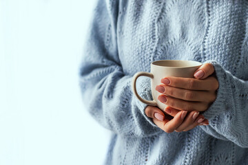 Female hands with a mug of drink close-up. Beautiful girl in a blue sweater holds a cup of tea or coffee in the morning sunlight. Autumn winter concept.