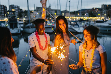 Obraz na płótnie Canvas Group of millennial friends celebrating with sparkles on a boat in harbour - Group of multiracial people having fun together at night party - Birthday celebration concept - Focus on african man