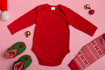 Mock up of a New Year's baby bodysuit with long sleeves in red on a pink background, decorated with gnome slippers, red pants and Christmas decorations, top view.